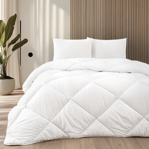 Ein weißes, rautenförmig gestepptes Bett mit zwei Kissen und Holz-Kopfteil in einem hellen Schlafzimmer mit Topfpflanze.
