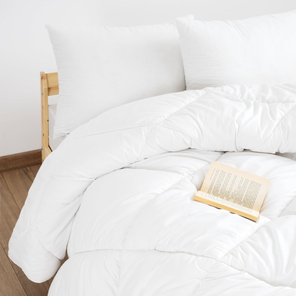 Ein weißes Bett mit zwei Kissen und einer 4-Jahreszeiten Mikrofaser Steppdecke. Ein offenes Buch liegt auf der hypoallergenen Decke.