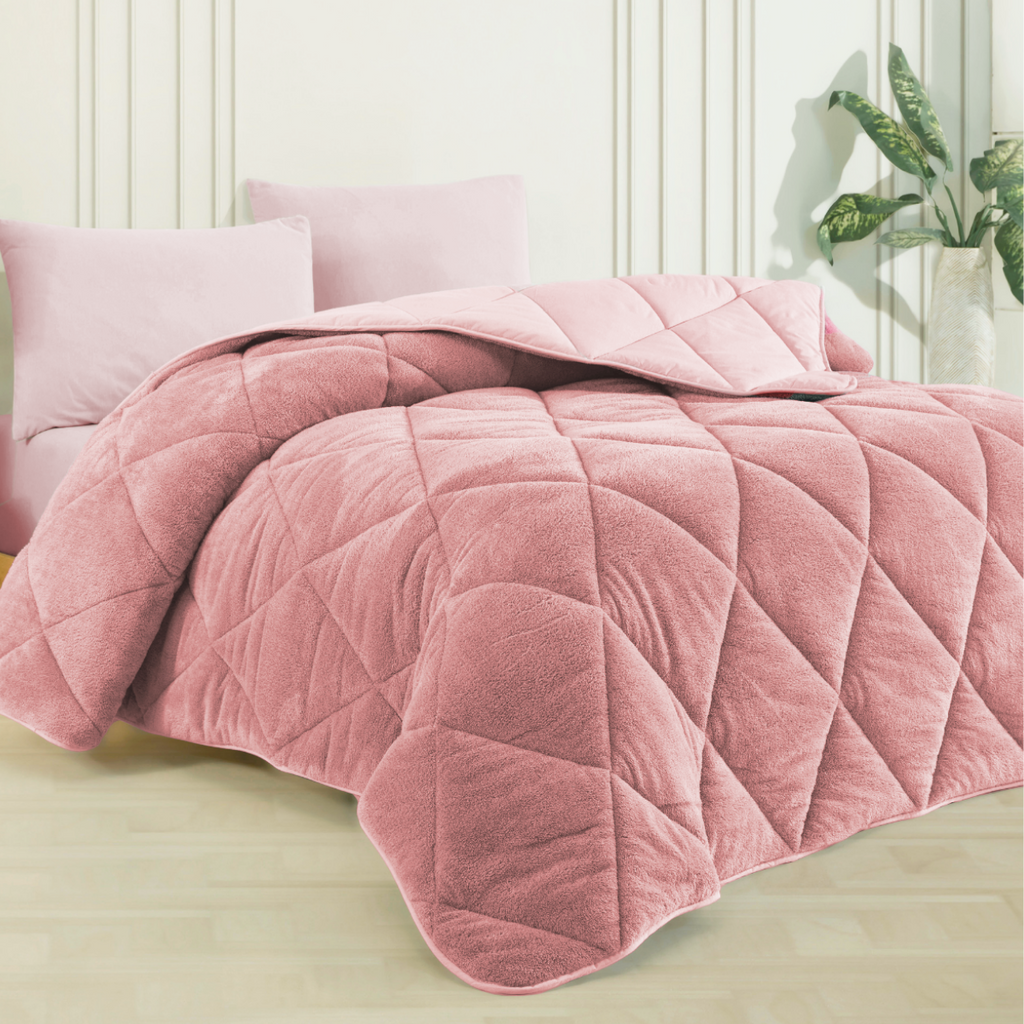Rosa Wellsoft-Steppdecke mit Rautenmuster auf einem Bett, dazu passende Kissen. Im Hintergrund eine weiße Wand und Pflanze.